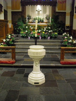 Stone alter steps with white marble baptismal font in front. There are flowers surrounding the alter and one the first step there is a baptismal candle. At the back of the alter there is an alter table with a cross hanging from the wall.