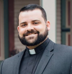 A white man with black hair wearing black clothes with a white clerical collar smiling.
