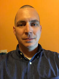 A white man with a shaved head wearing a blue shirt smiling.