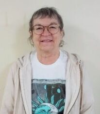 A white woman with gray hair and glasses wearing a white T-shirt smiling.